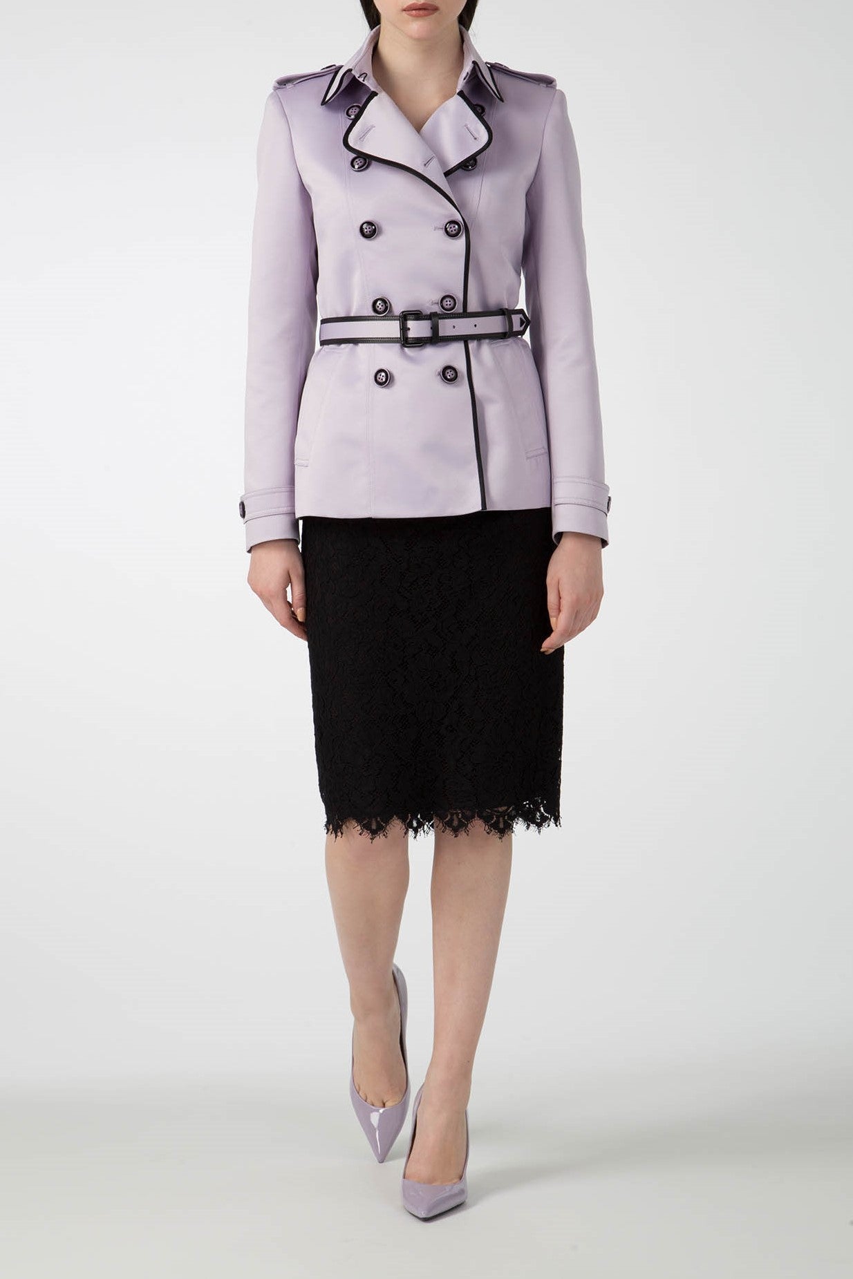 A person wearing an Antonio Croce Taylor Trench Coat with Belt in a vintage double-breasted lavender style and black trim, paired with a black lace skirt and matching lavender heels, stands against a plain white background.